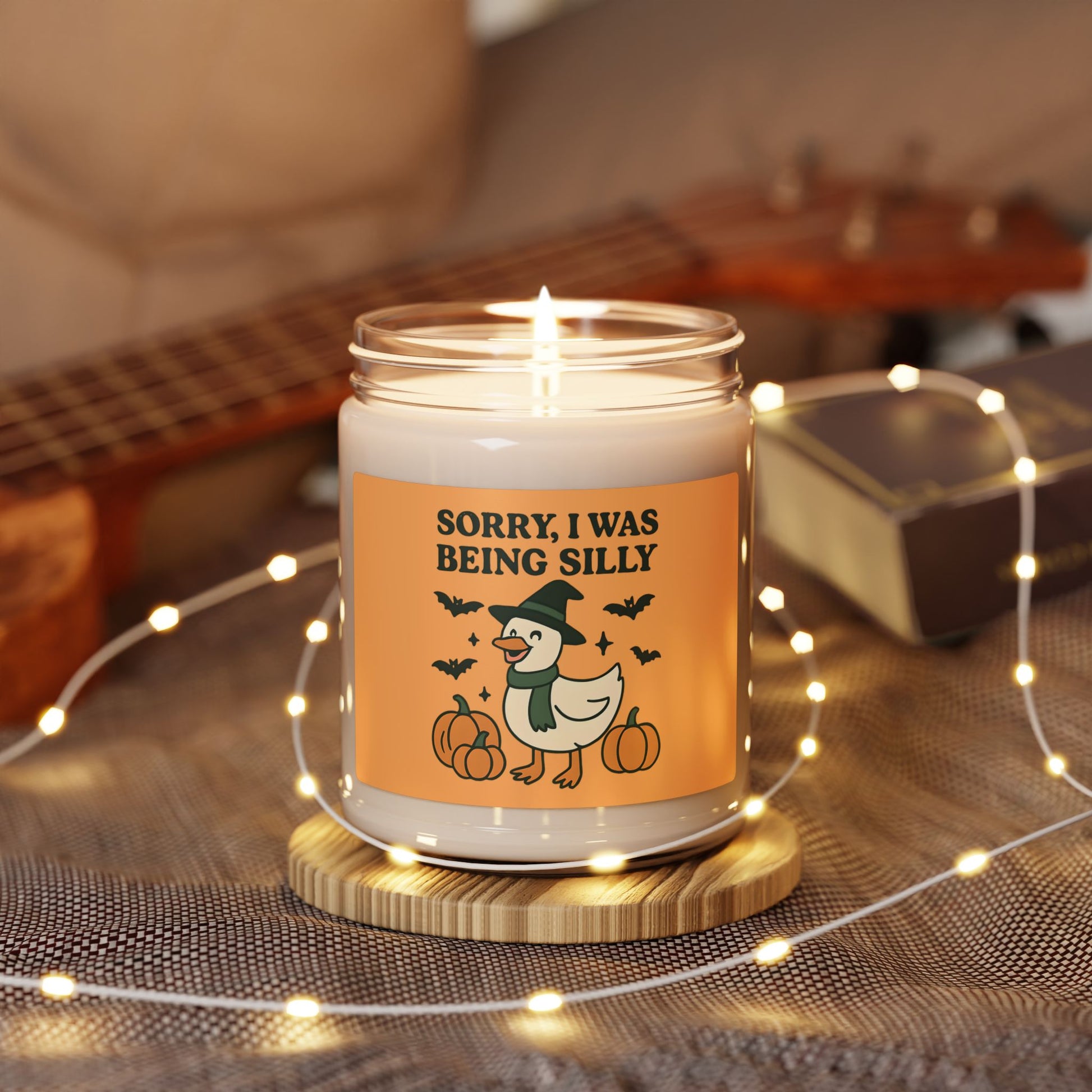 Candle with a Halloween-themed label on a wooden coaster, surrounded by string lights and a guitar in the background.