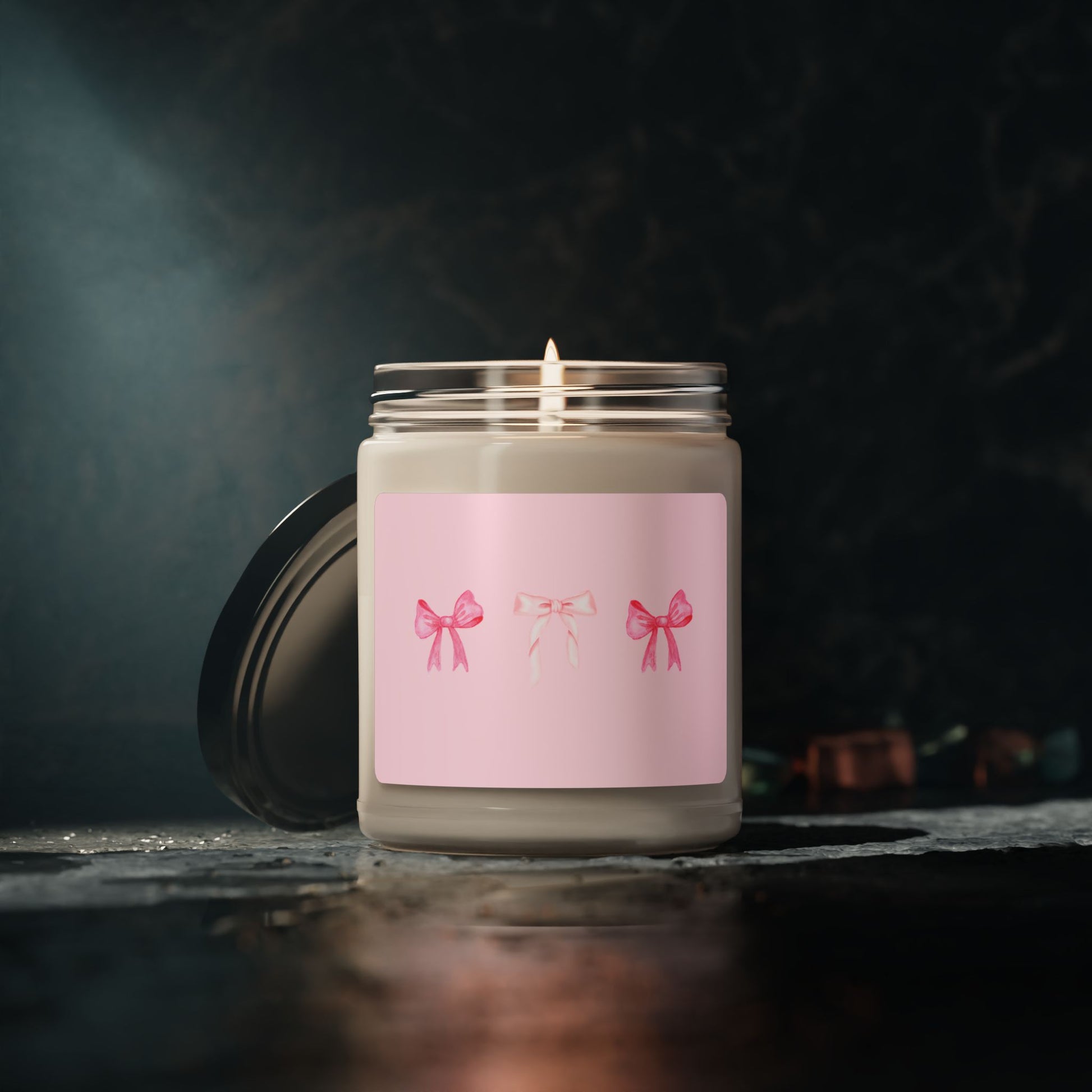 Candle in a glass jar with a pink label featuring three bows on a dark background
