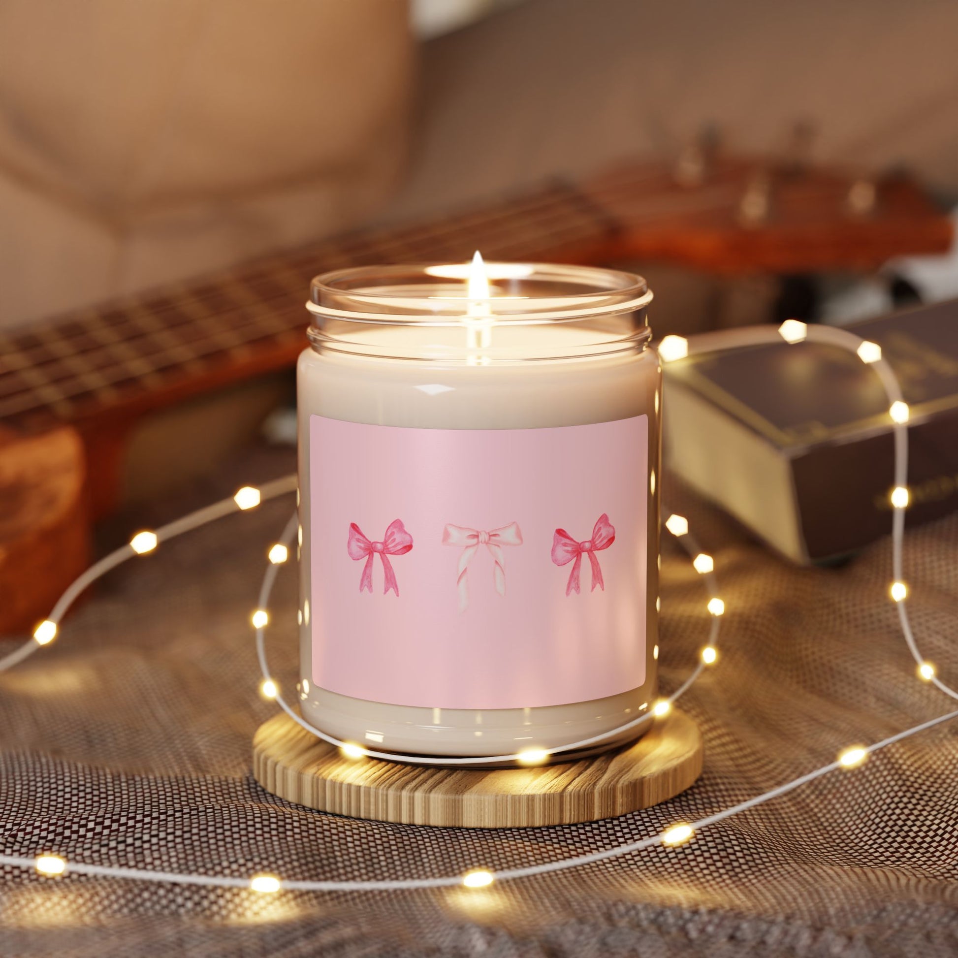 Candle with a pink label featuring bows on a wooden coaster with string lights and a guitar in the background.